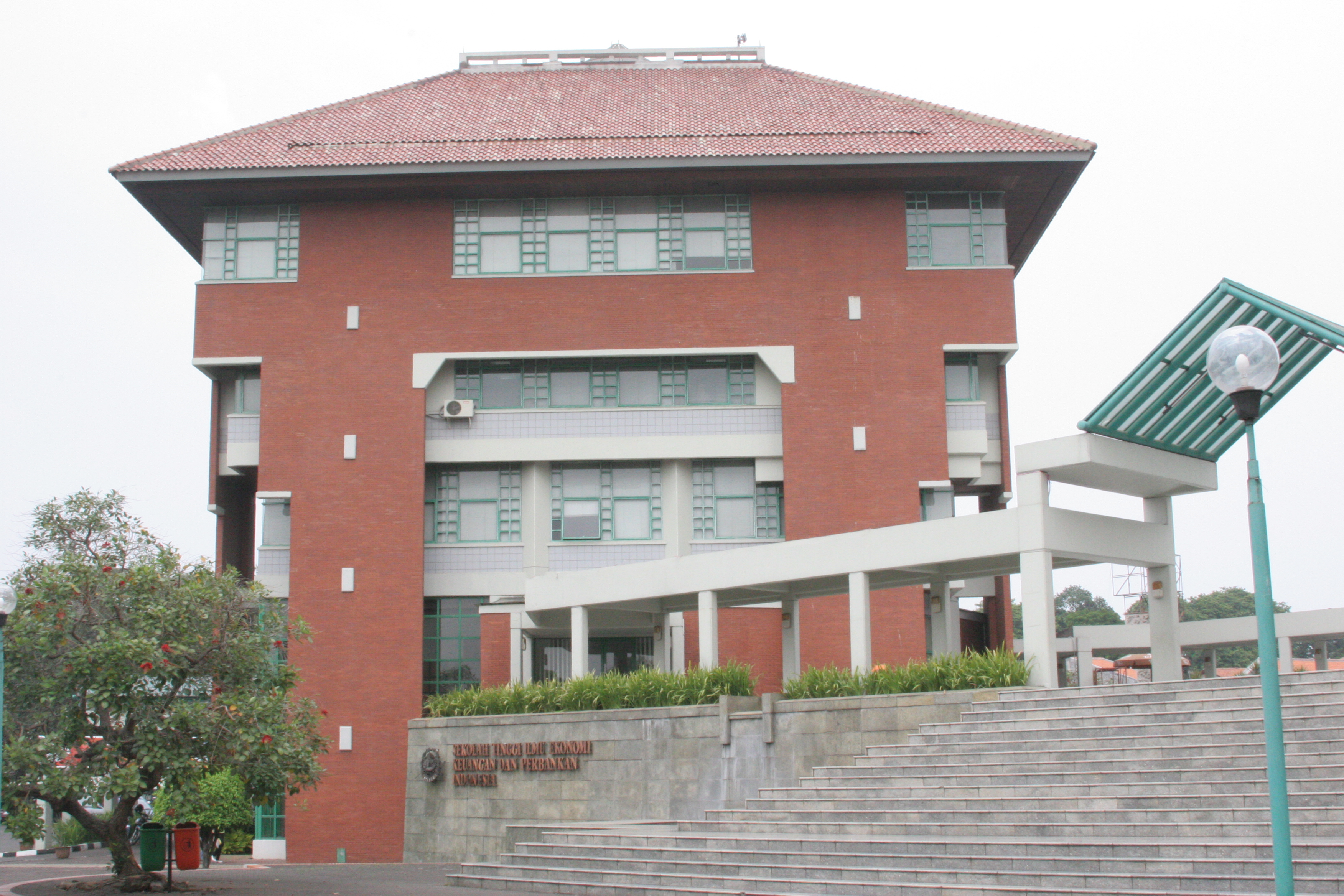Gedung rektor Universitas Trilogi berwarna merah dengan atap dan jendela berbingkai hijau dan garis putih di lantai tiga dan empat. Berdiri dengan plaza di depannya.
